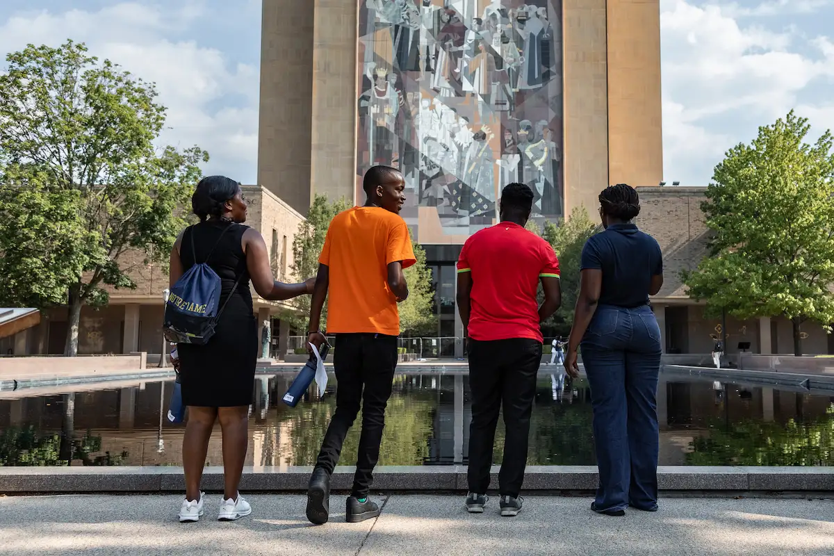 Four students stand facing the Hesburgh Library's Word of Life mural. A woman in a black dress with a blue Notre Dame backpack, a smiling man in an orange shirt, a man in a red shirt, and a woman in a navy polo and jeans. The reflecting pool is visible below.