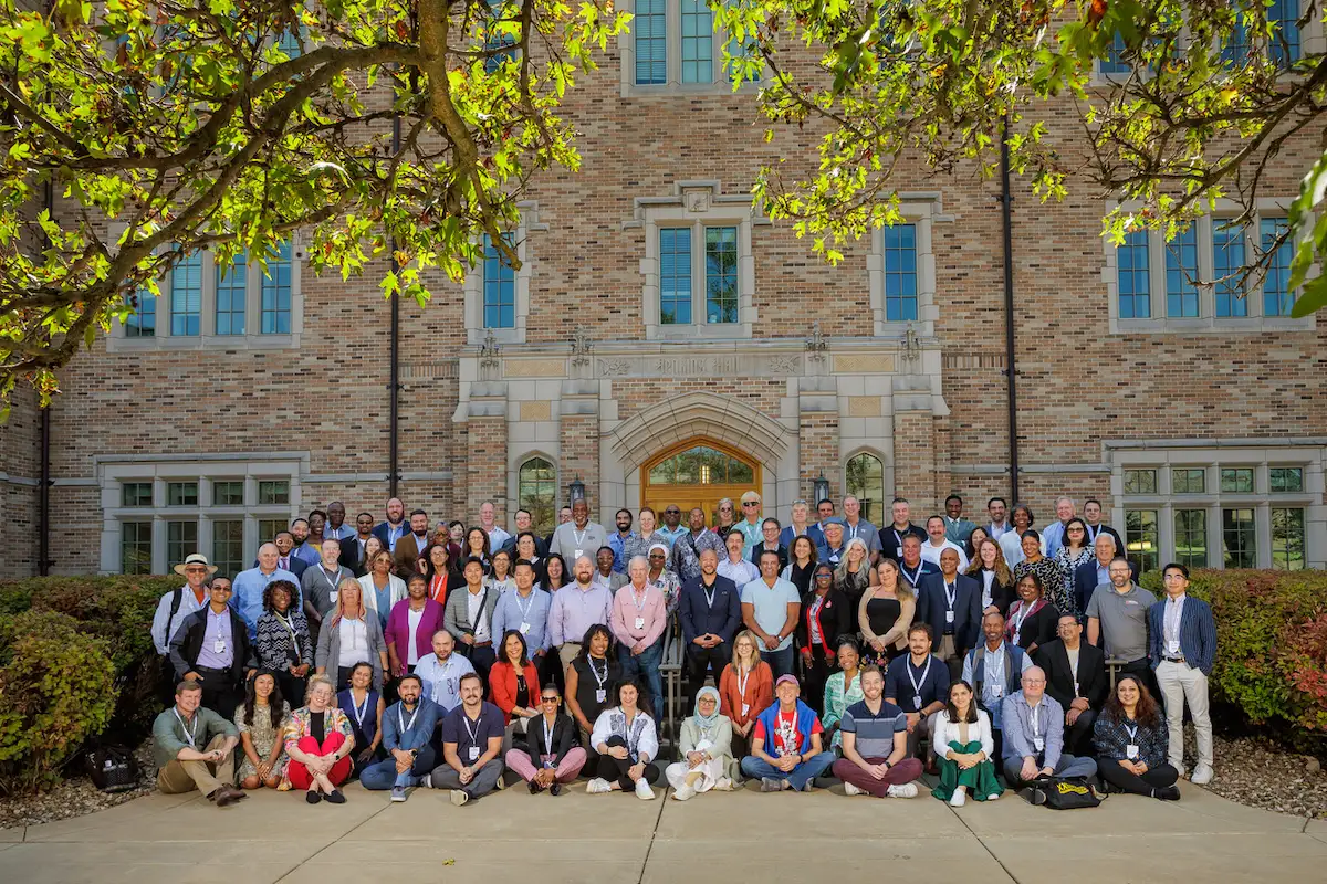Many diverse people, some seated and some standing, smile for a group photo in front of Notre Dame's Prima Hall, a brick building with a stone arched entryway.