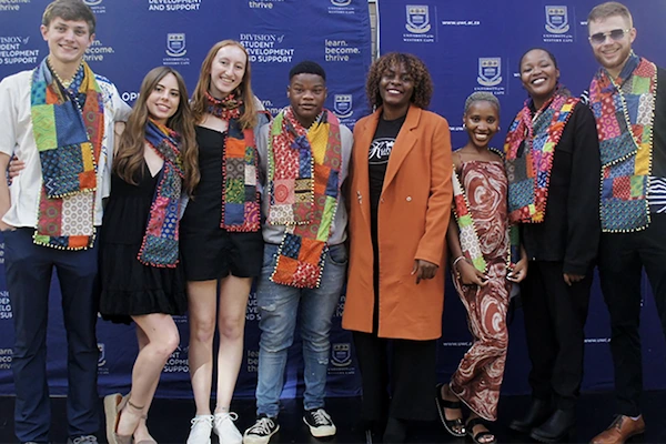 Eight diverse young adults smile, wearing vibrant patchwork scarves against a blue University of Western Cape banner that reads 'learn. become. thrive.'