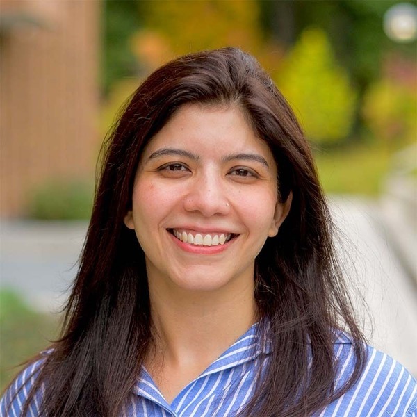 Smiling woman with dark hair and a nose piercing, wearing a blue and white striped shirt, with blurred foliage behind her.